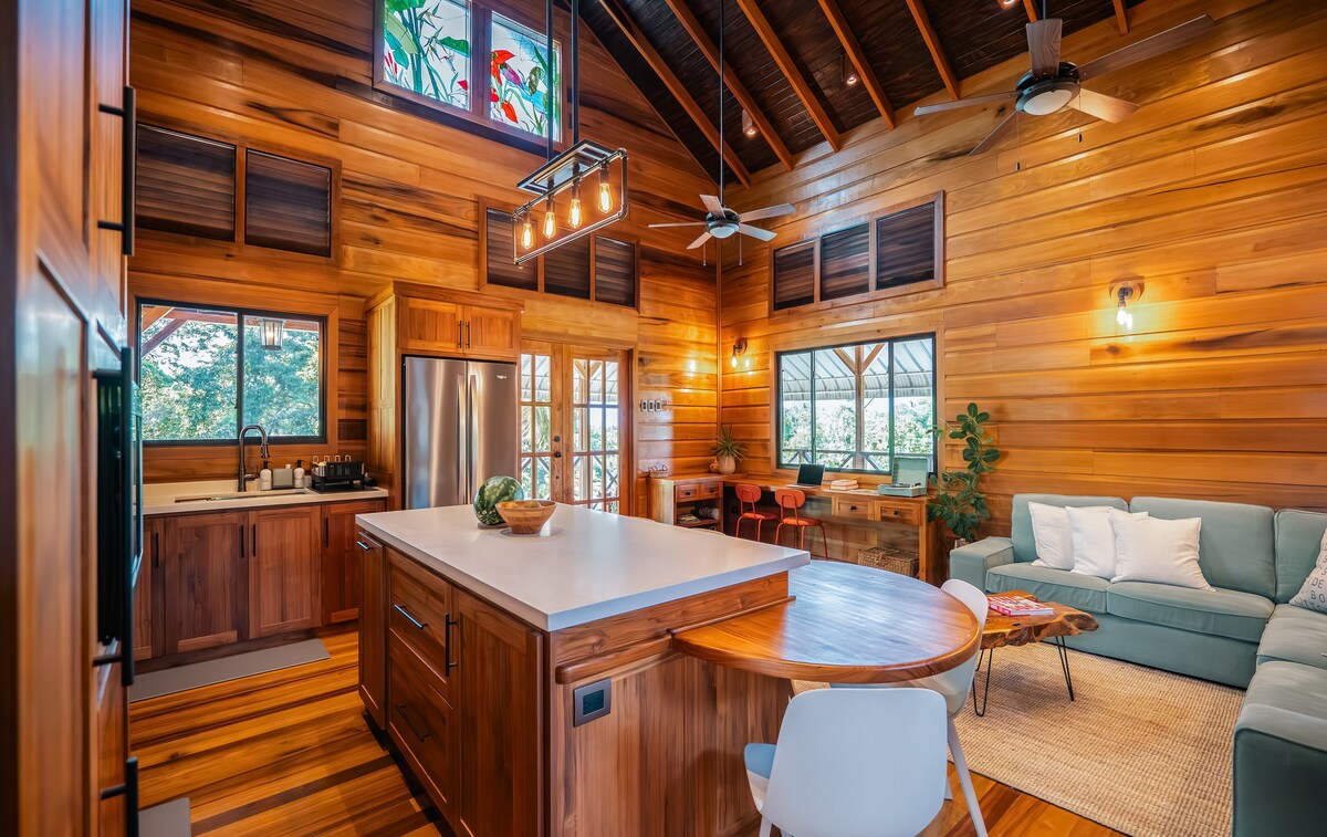 An open-concept kitchen and living area features wooden walls and ceiling beams, enhancing a warm atmosphere. A central island with a light countertop is surrounded by natural wood cabinetry. Windows allow natural light to illuminate the space, while a comfortable seating area with a sofa is visible.
