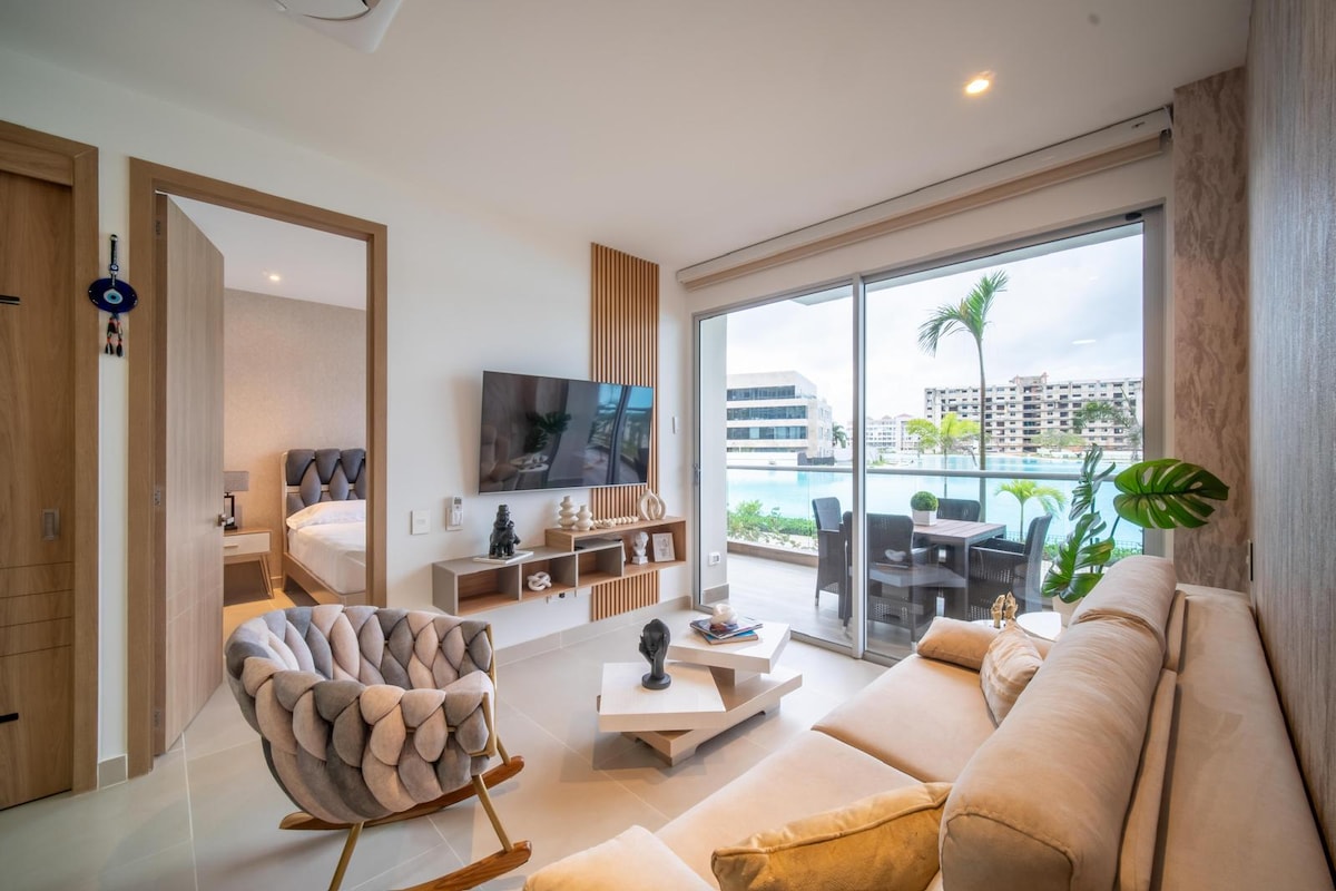 A modern living area features a spacious beige sofa, a sleek coffee table, and a large wall-mounted TV. Floor-to-ceiling glass doors open to a view of the lagoon, allowing natural light to fill the room. The space connects to a bedroom through an open doorway.