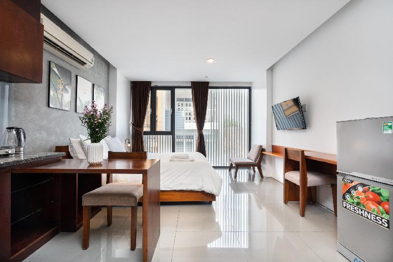 A modern room features a spacious bed and a small dining area with a table and chairs. Large windows allow natural light to flow in, with sheer curtains providing privacy. A flat-screen TV is mounted on the wall, and a small refrigerator is present.