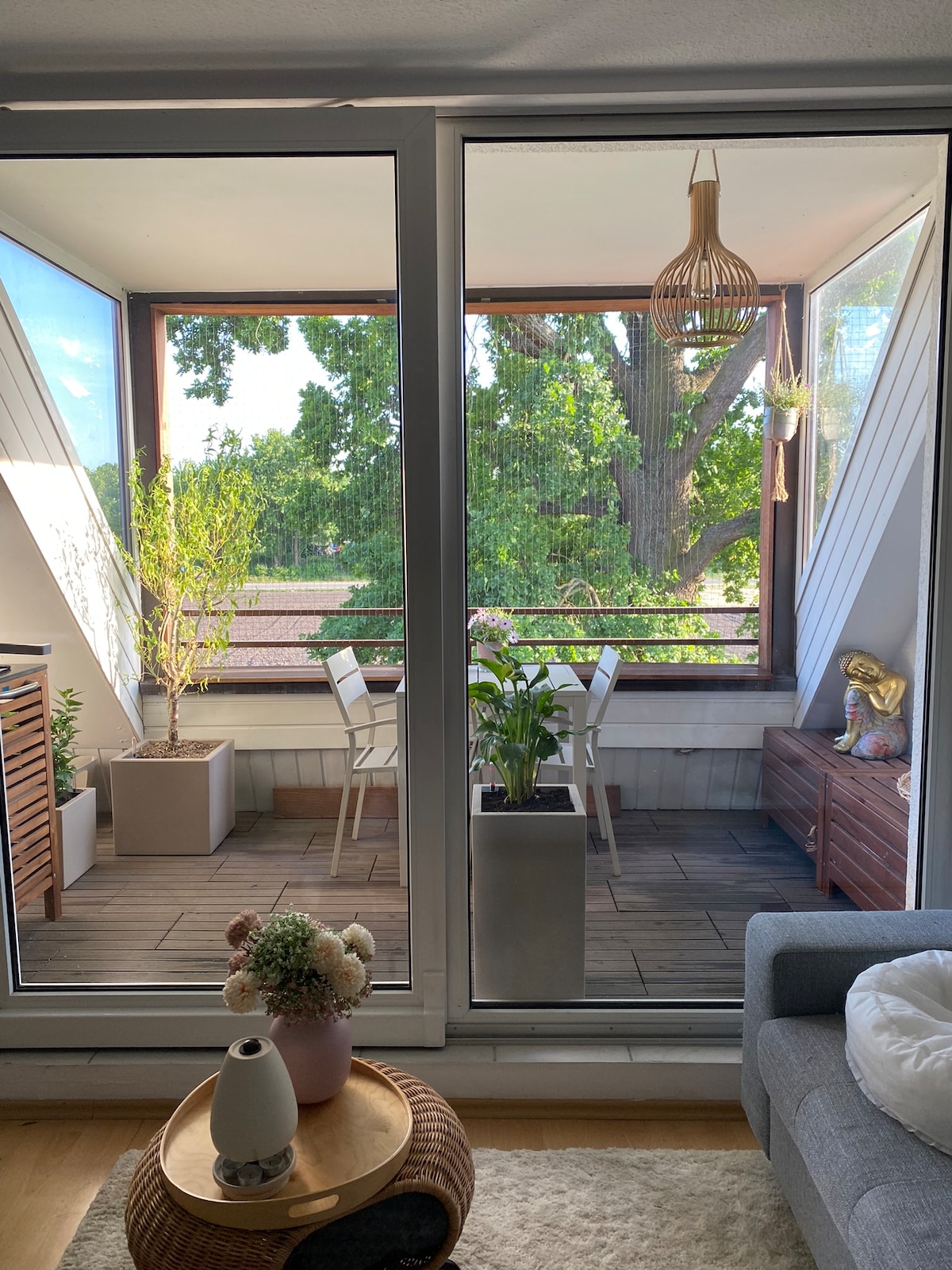 A balcony area is visible through large glass doors, featuring a small dining table with two chairs surrounded by greenery. Planters and decor add a natural touch. A comfortable seating area is seen in the foreground, enhancing the indoor-outdoor connection.