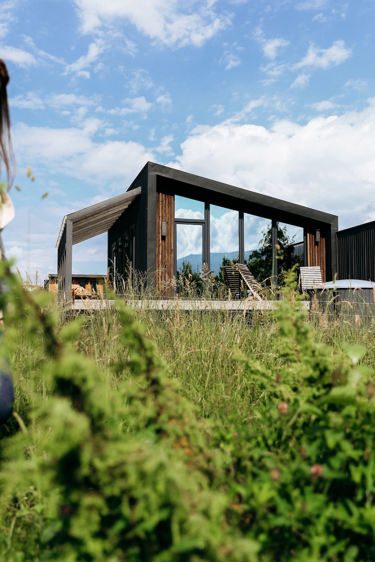 A modern villa is set against a backdrop of blue skies and wispy clouds. The structure features large glass windows and wooden accents, allowing for ample natural light. Surrounding wild grass adds to the serene outdoor setting of the property.