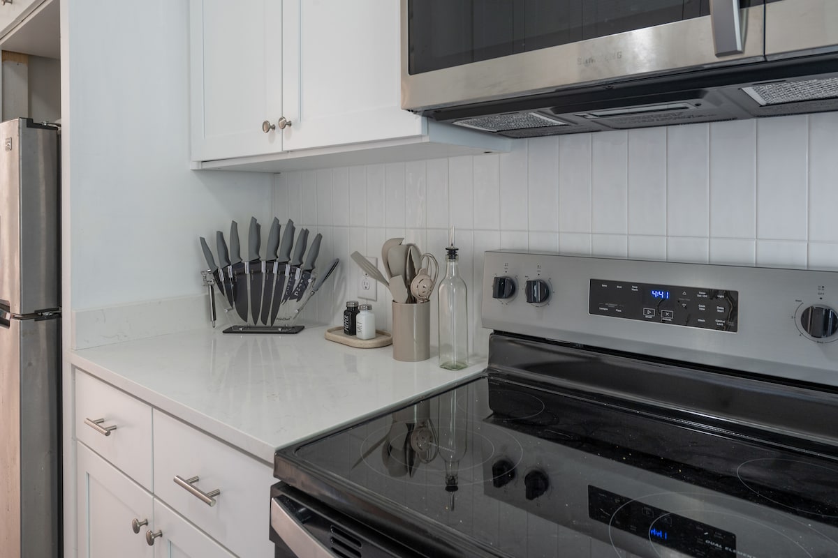 The newly designed kitchen showcases sleek stainless steel appliances and a smooth countertop. A collection of kitchen knives is neatly arranged on a block, while an array of essential cooking tools is visible. Modern cabinetry adds to the overall contemporary design.