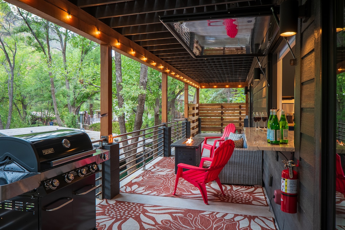 A spacious deck features comfortable seating with bright red chairs, placed on a patterned outdoor rug. A modern barbecue grill is situated nearby, under warm ambient lighting provided by string lights above. Lush green trees frame the serene view, enhancing the inviting outdoor atmosphere.