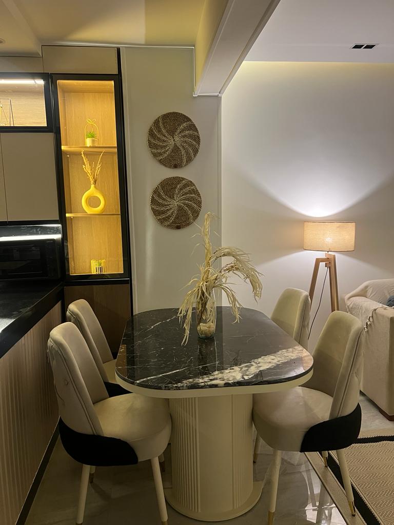 A modern dining area features a round marble table surrounded by four upholstered chairs. Decorative wall art made from natural materials adds texture, while warm lighting from a nearby lamp creates a cozy ambiance.