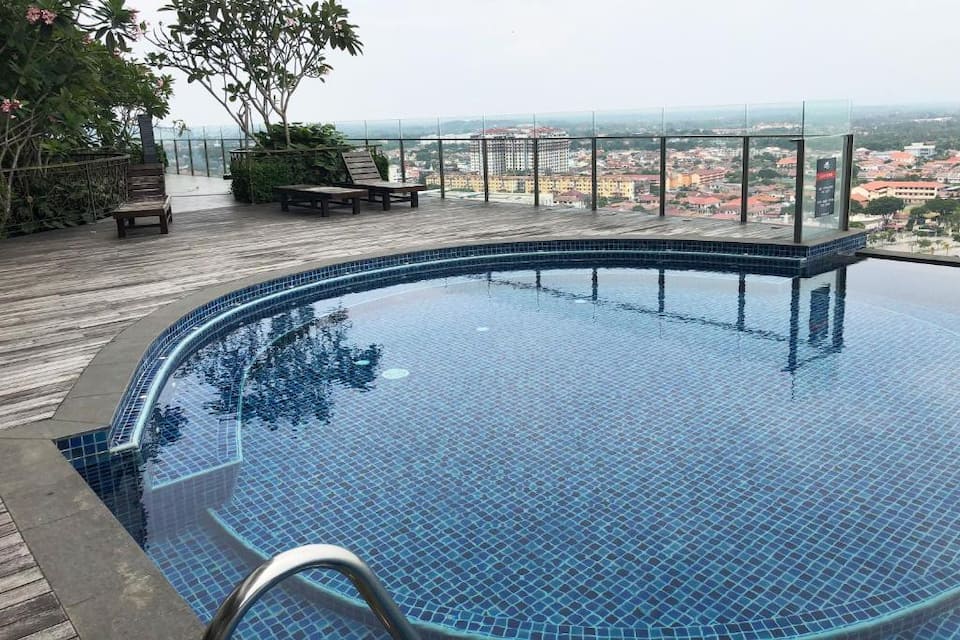 An elevated pool area is shown, featuring a circular pool with vibrant blue tiles. Lounge chairs are positioned nearby, surrounded by lush greenery and glass railings offering expansive views of the surrounding cityscape.