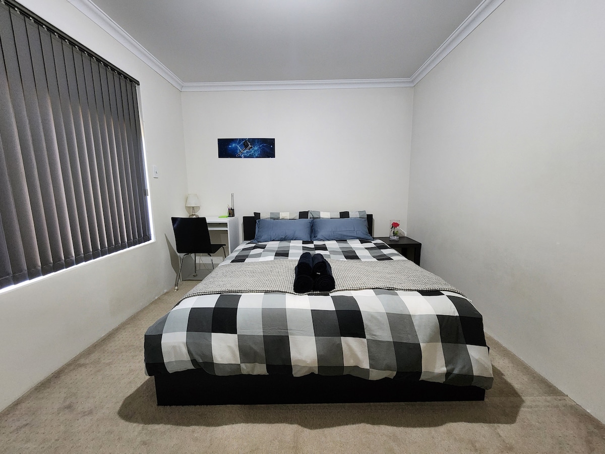 A spacious sleeping area features a large, checkered bedspread covering a comfortable bed. A neutral-colored wall complements the room, while a desk and chair are positioned near the window, which is dressed with vertical blinds. Soft towels are neatly arranged on the bed.