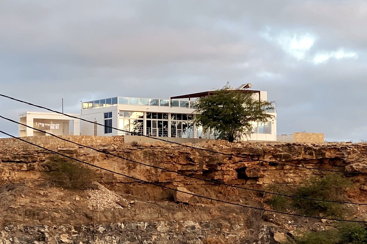A modern penthouse is perched on a rocky cliff, showcasing large glass windows that reflect the surrounding landscape. The structure is designed with clean lines, and its elevated position provides a sense of openness to the horizon.