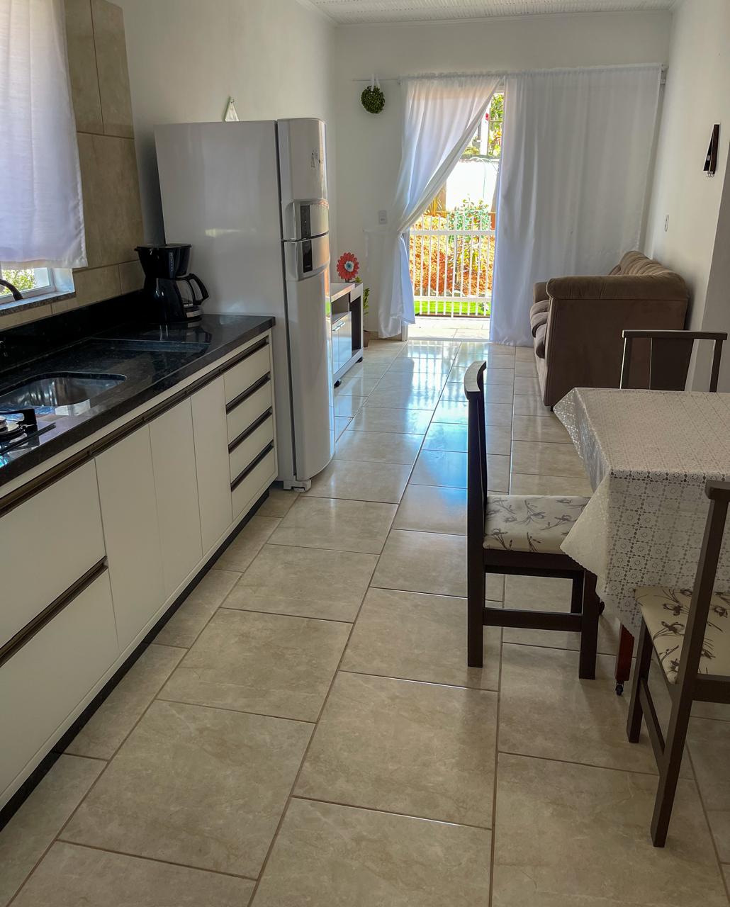 A modern kitchen area features sleek cabinetry and a large refrigerator. A coffee maker rests on the counter beside a inviting dining table with four chairs. Natural light filters in through a window, illuminating the open space adjoining a comfortable living area with a couch.