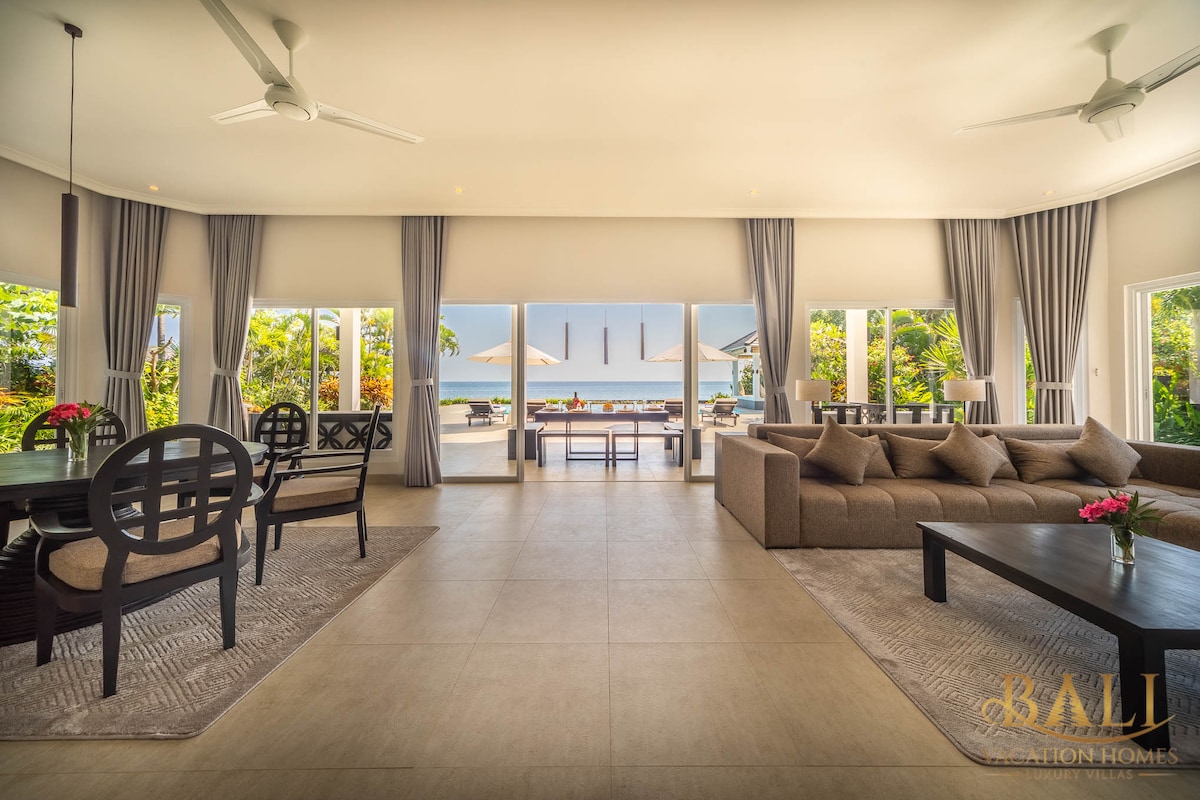 An open-concept living space features a dining area with black chairs and a large wooden table, along with a cozy seating area with a sectional sofa. Large glass doors provide a view of the pool terrace and Bali Sea, creating a fluid connection between indoor and outdoor living.