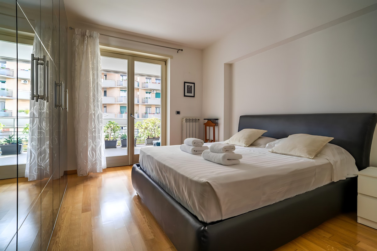 A spacious bedroom is featured with a large bed, dressed in soft white linens and adorned with two decorative pillows. Natural light pours in through large windows, which overlook a balcony with plants. A mirrored wardrobe and bedside tables complete the calming space.