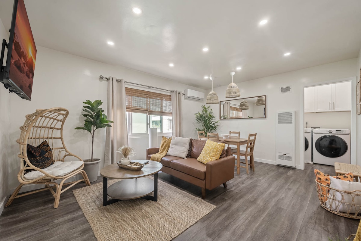 A spacious living area features a comfortable seating arrangement with a dark coffee table at its center. A wall-mounted TV is visible opposite a stylish dining table. Natural light fills the space, highlighting the modern decor and greenery, while a washer and dryer are tucked against one wall.