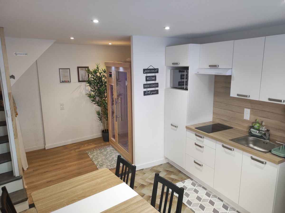 A modern kitchen is displayed with sleek white cabinets and wooden accents. A dining table with black chairs is positioned nearby. Natural light brightens the space, enhancing the inviting atmosphere. A sauna area is visible in the background, accessed through a partially open door.