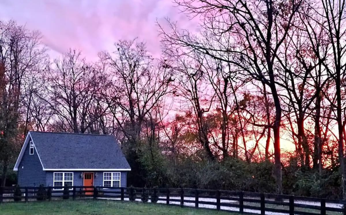A cozy cottage is set against a vibrant sunset, with soft hues of pink and orange in the sky. The property is surrounded by bare trees and a black wooden fence, enhancing the serene rural atmosphere.