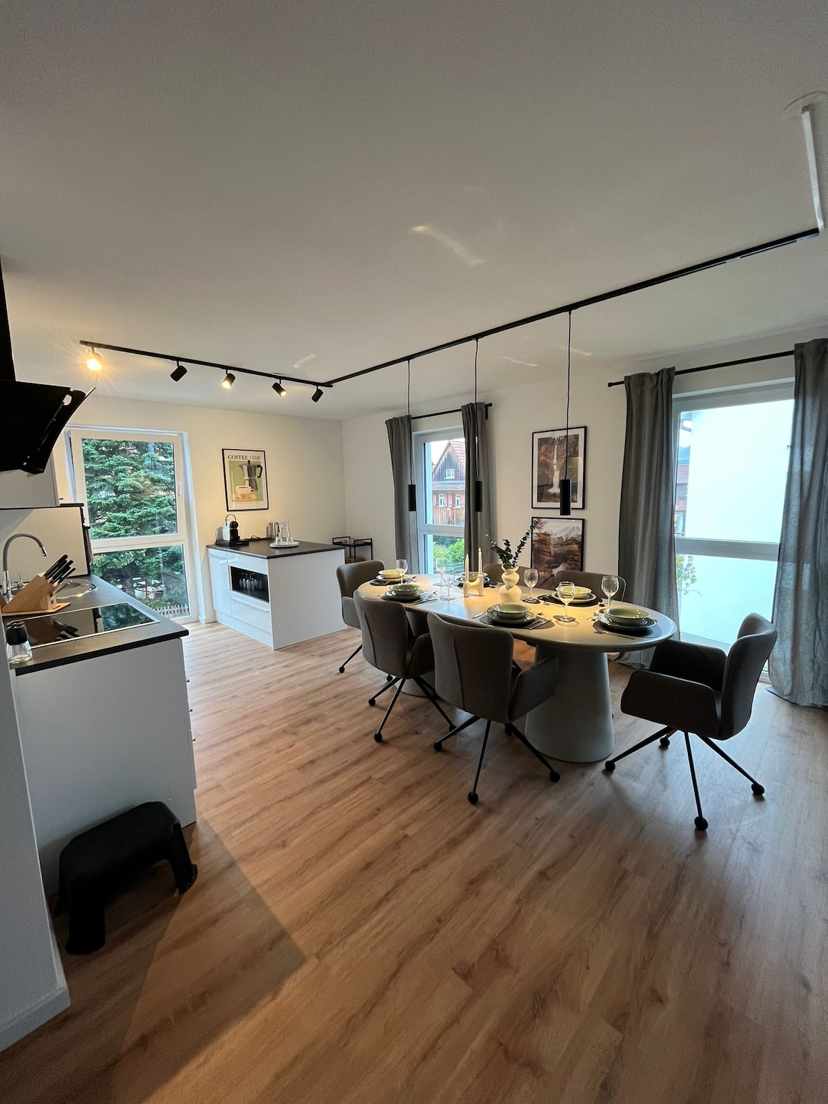 A modern kitchen and dining area features a round dining table set for six, surrounded by comfortable chairs. A sleek kitchen with a stovetop and oven is visible in the background, complemented by large windows that provide natural light and views of the outdoor greenery.