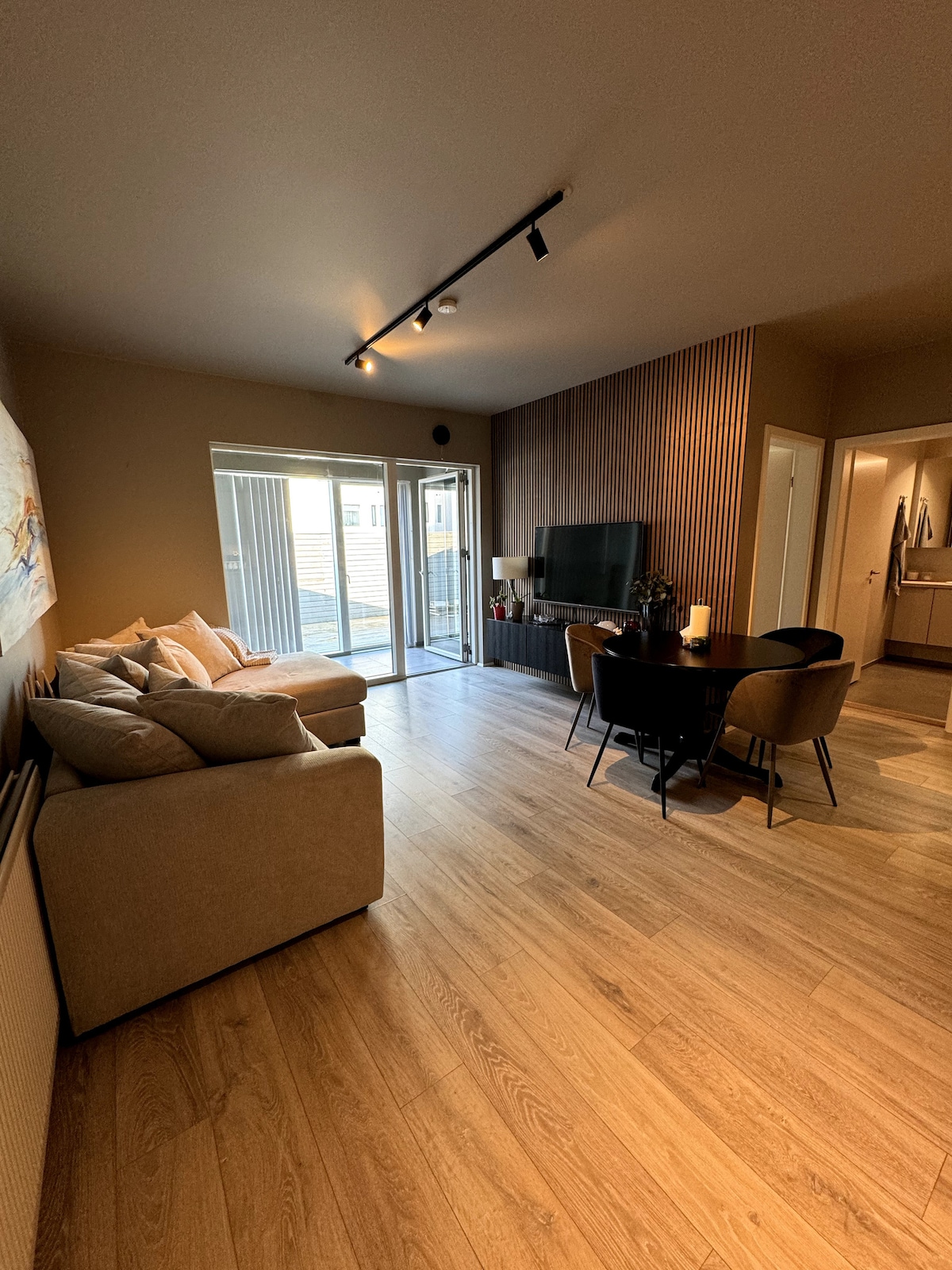A spacious living area features a comfortable sofa and a dining table with black chairs. Large sliding glass doors provide access to an outdoor space, while a television is mounted on the wooden accent wall. Warm lighting enhances the inviting atmosphere.