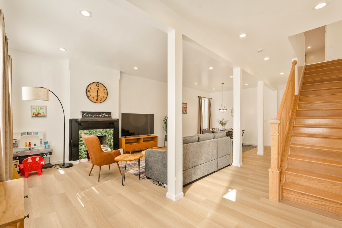 A bright, open-concept living area showcases a comfortable gray sofa and a mid-century style chair facing a modern television. Natural light floods the space, complemented by a large wall clock and a decorative fireplace. The wooden staircase adds warmth to the inviting atmosphere.