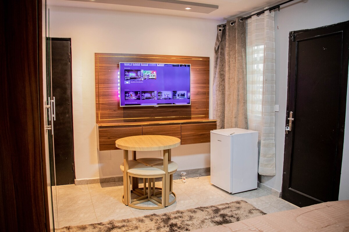 A compact living space features a round dining table paired with three chairs. A flat-screen television is mounted on a wooden accent wall, with a small white refrigerator situated nearby. Natural light enters through a window with sheer curtains.