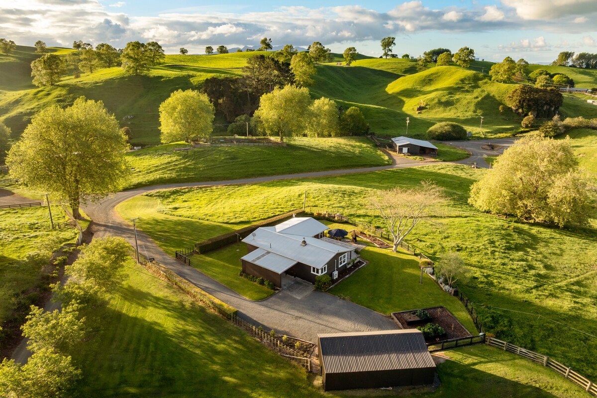 Aerial view of a renovated cottage surrounded by lush green hills and scattered trees. A winding driveway leads to the cottage, with grassy areas providing ample outdoor space. The setting reflects a serene rural landscape, showcasing the peacefulness of the location.