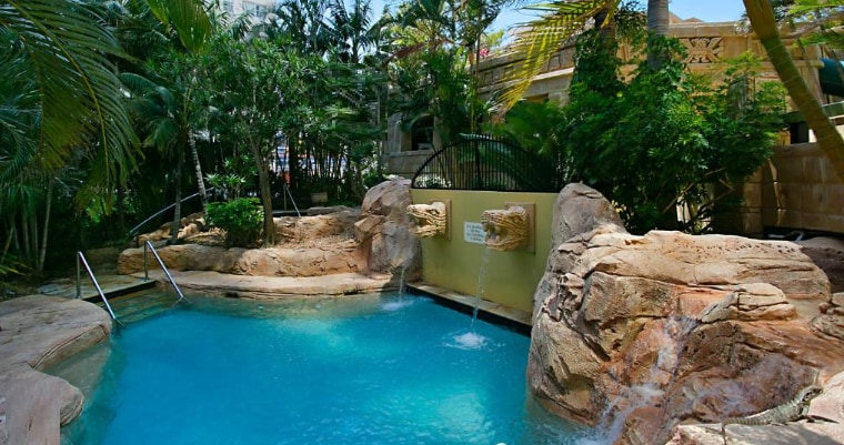 A serene pool area is surrounded by lush tropical plants and textured rock formations. Water cascades gently from stone features into the clear blue water, inviting relaxation. A set of metal stairs leads into the pool, providing easy access.
