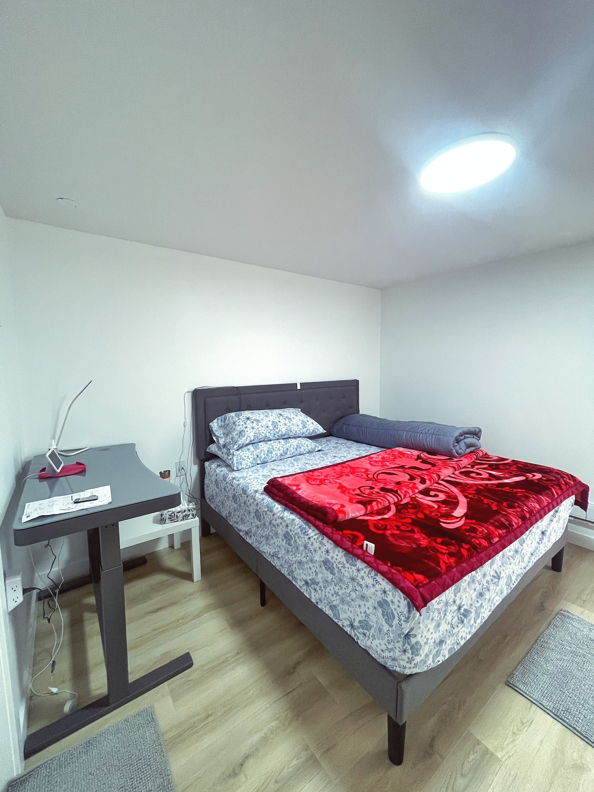 A cozy sleeping area is presented, featuring a padded bed topped with a floral-patterned duvet and a red throw blanket. A compact desk with a lamp is positioned beside the bed, while soft area rugs cover the floor in a well-lit space with white walls.