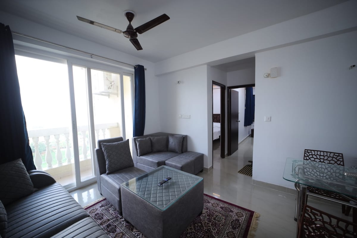 A modern living area features a comfortable gray sofa set with plush cushions, accompanied by a glass coffee table. Natural light floods through large sliding glass doors, which lead to a balcony. A dining table with intricate detailing is positioned nearby, while a doorway leads to additional sleeping areas.