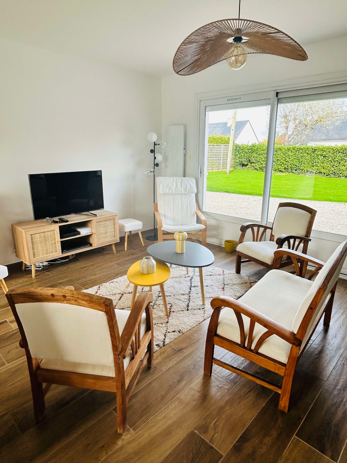A bright living area contains a flat-screen television and a wooden media console. Four chairs surround a small coffee table, while a plush area rug adds texture to the floor. Large sliding glass doors open to a well-maintained garden, inviting natural light into the space.