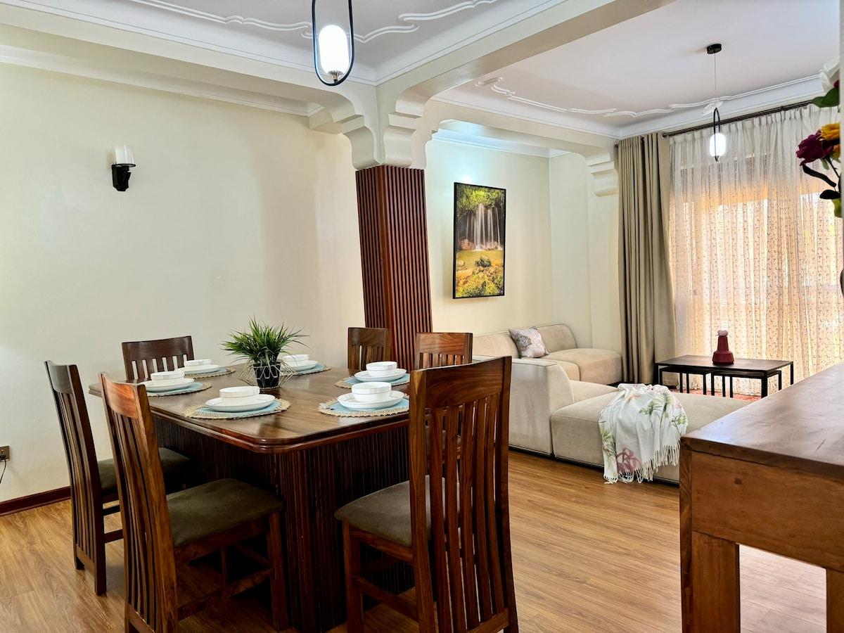 The dining area features a wooden table set for six, surrounded by matching chairs. A light-colored couch is positioned nearby, with a decorative painting reflecting nature decor on the wall. Soft natural light filters through sheer curtains, creating a warm ambiance.