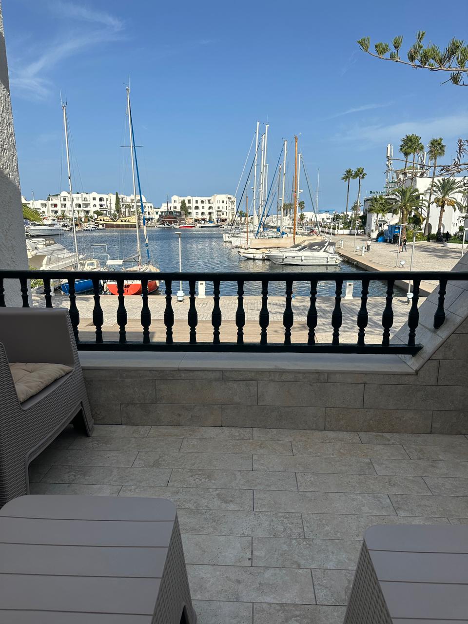 A spacious terrace is framed by wrought iron railings, offering a view of the marina filled with sailboats. Bright blue skies and palm trees provide a scenic backdrop to the calm water, inviting relaxation in the outdoor seating area.