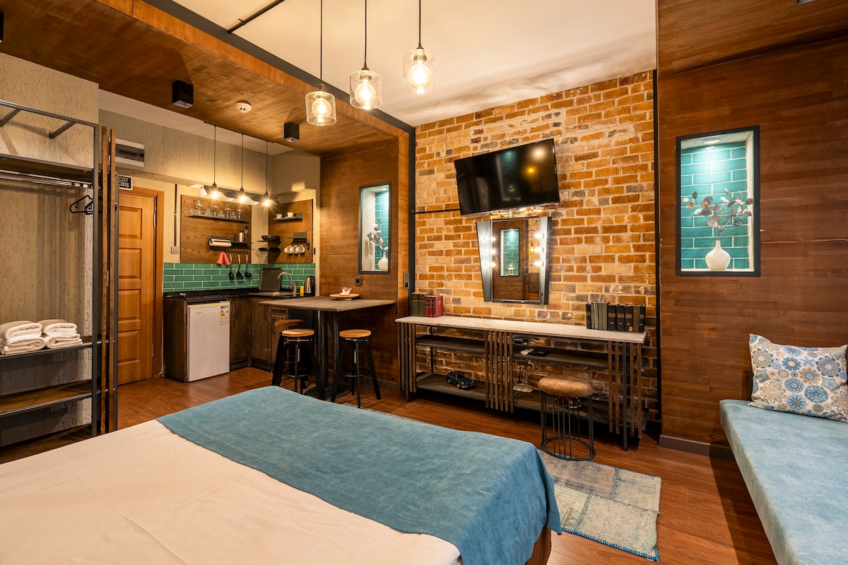 A modern room showcases a spacious bed covered with a light blue blanket. A rustic brick wall features shelves with glassware and kitchen necessities. A small kitchenette is equipped with a refrigerator. Warm lighting enhances the inviting space, complemented by a stylish seating area.