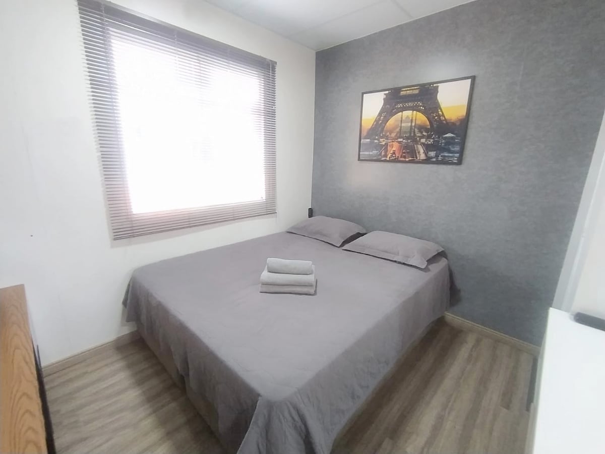 A cozy sleeping area features a spacious bed with gray bedding and two neatly folded towels on top. Natural light enters through the window, framed by light-colored blinds. A framed image of the Eiffel Tower adds a decorative touch to the room's neutral walls.