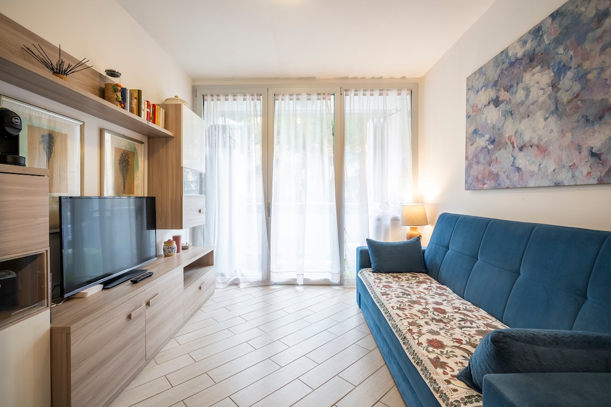 A comfortable living area features a blue sofa with a decorative throw and a sleek wooden media unit. A television is positioned on the unit, while natural light filters through sheer curtains, illuminating the light-colored tiled flooring and soft wall art.