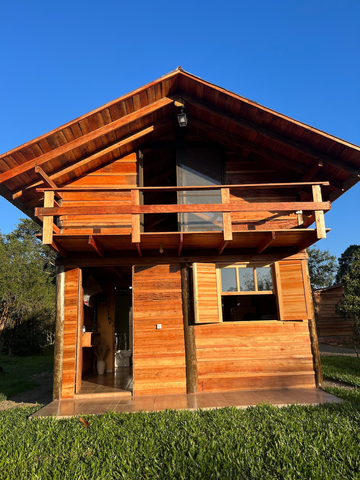 The exterior of a rustic wooden cabin features a sloped roof and a spacious balcony. Large windows allow natural light to enter, while the surrounding lush green grass enhances the cabin's connection with nature.