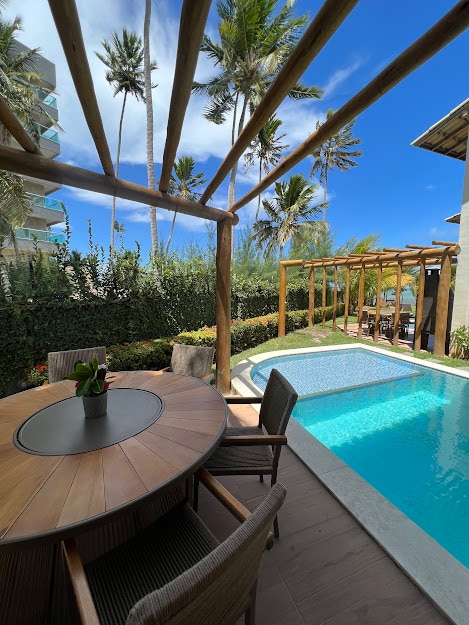 A wooden dining table with several chairs is positioned under a trellis, providing shade by the pool area. A private pool reflects the blue sky, surrounded by lush greenery and palm trees. The outdoor space offers a seamless integration with nature.
