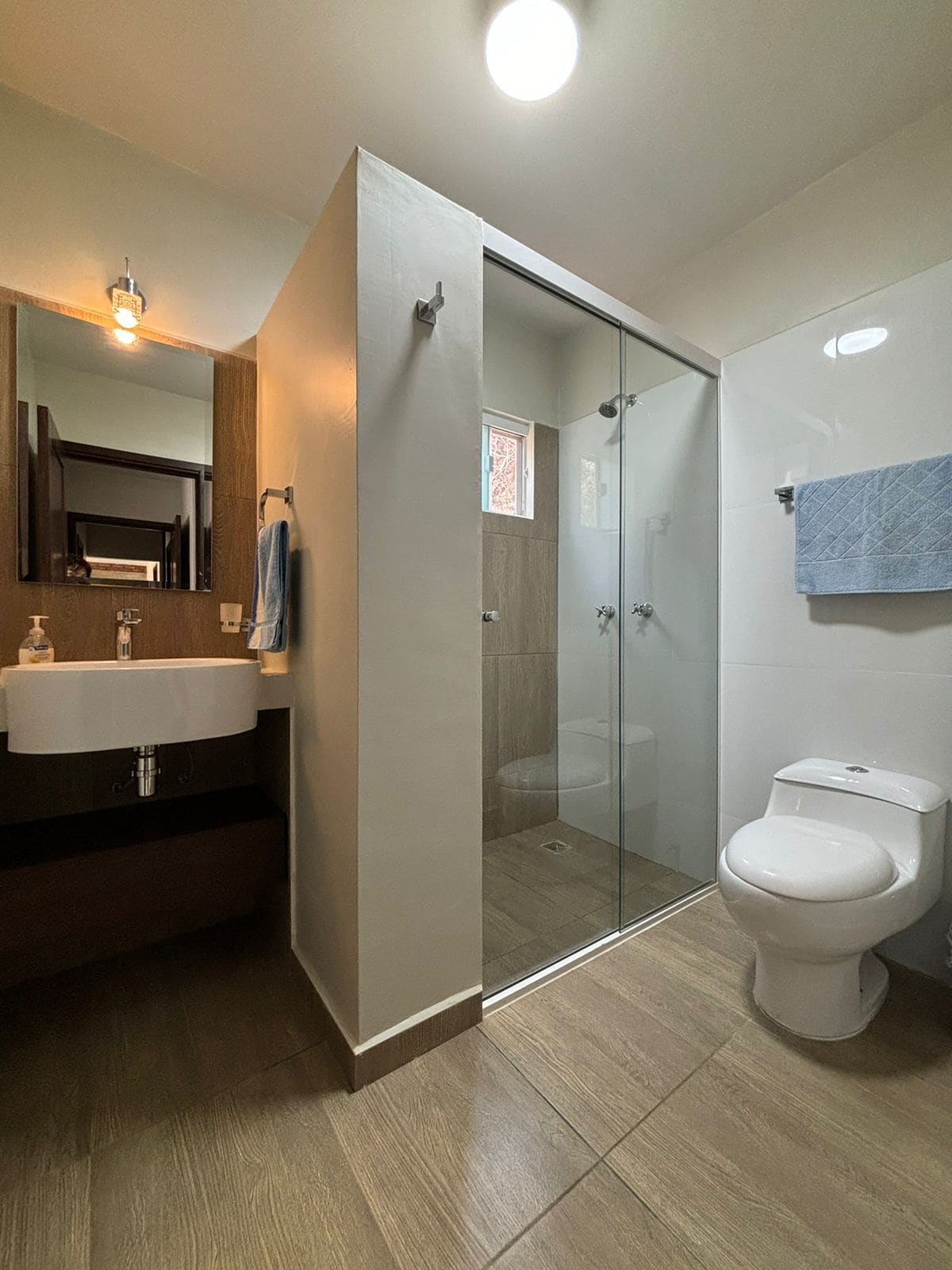 A clean and modern bathroom is depicted, featuring a glass shower enclosure and a compact sink mounted on a wooden vanity. Light fixtures enhance the space's brightness, while a soft blue towel hangs on the wall. Neutral-toned tiles cover the floor and walls.