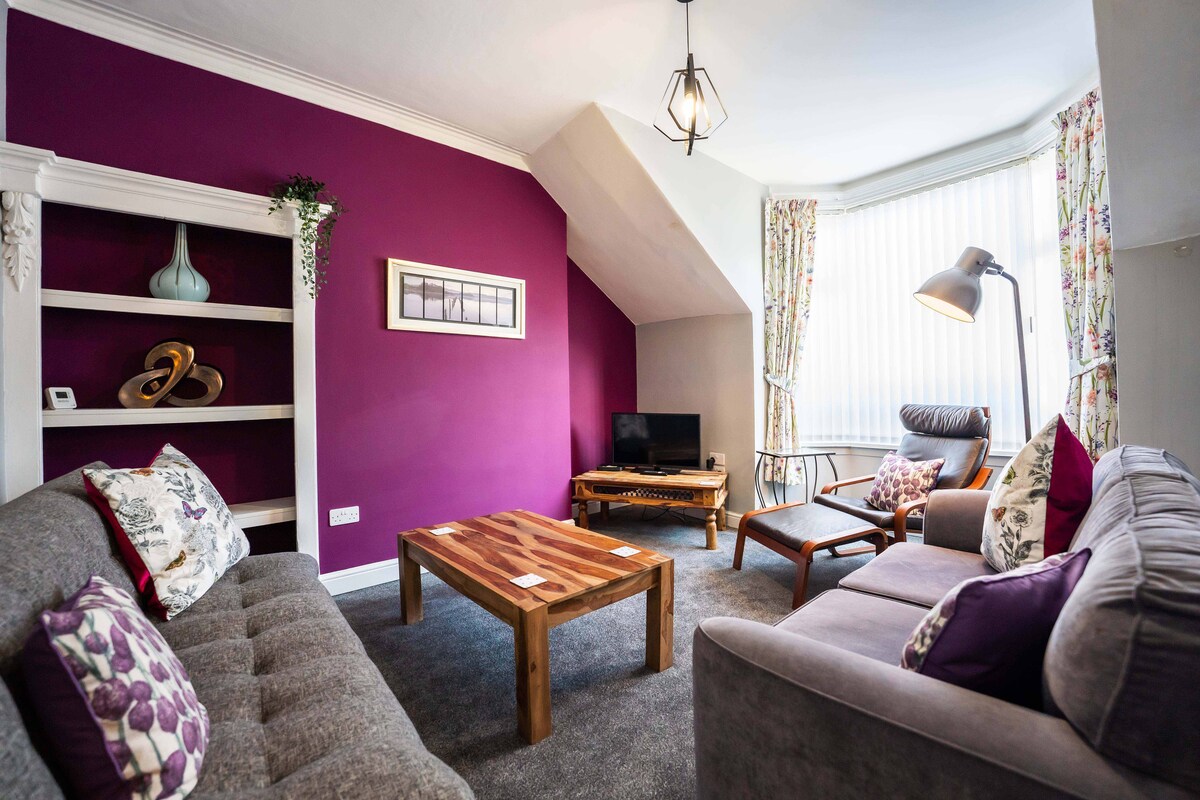 A spacious lounge area features a mix of comfortable seating, including a gray sofa and wooden armchair. A small wooden coffee table is positioned in the center. A smart TV is placed on a shelf against a bright purple wall adorned with decorative elements and framed artwork.