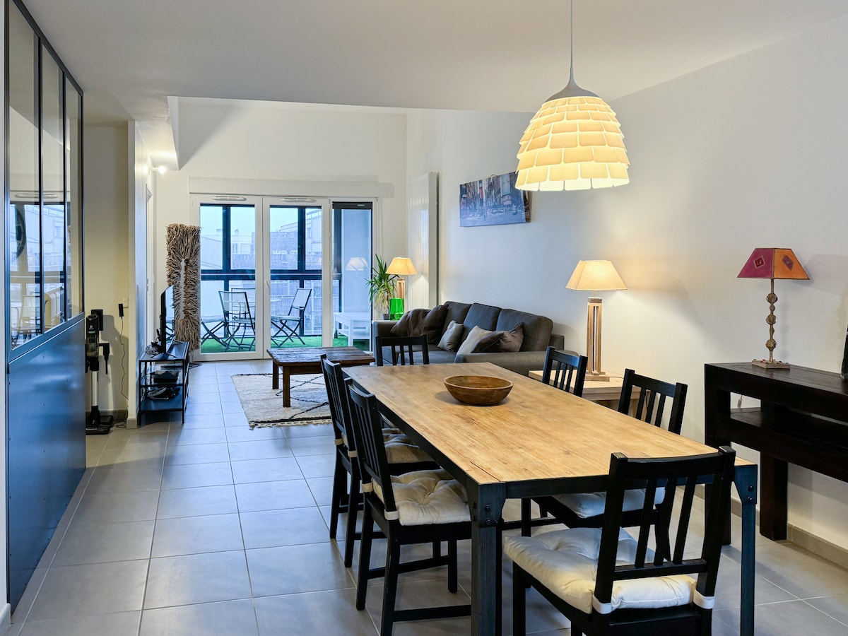 An open living space features a dining table with black wooden chairs and a light wooden top. A comfortable couch is positioned nearby, accompanied by two side lamps. Large glass doors lead to a balcony, allowing natural light to fill the room. A soft rug adds warmth.