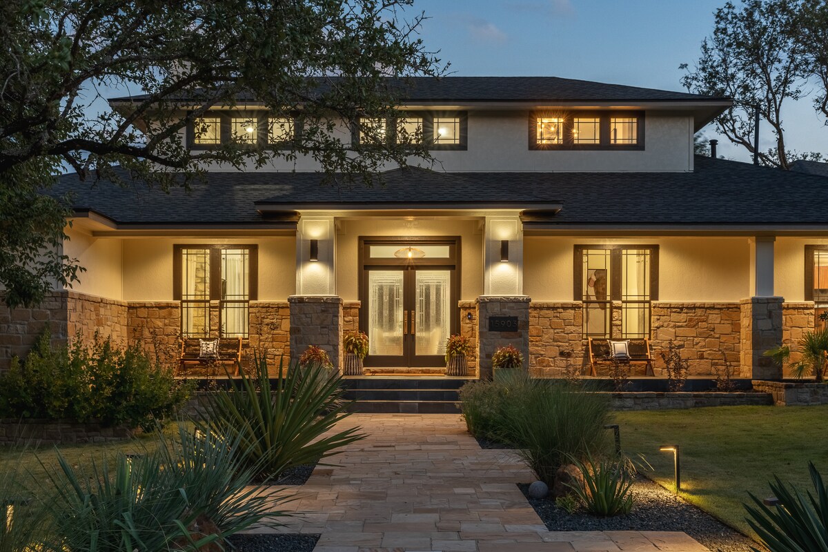 The contemporary retreat is lit warmly at dusk, showcasing a welcoming entrance framed by two rocking chairs. Stone accents complement the modern facade, while well-manicured landscaping leads to the front door, creating an inviting setting for guests.