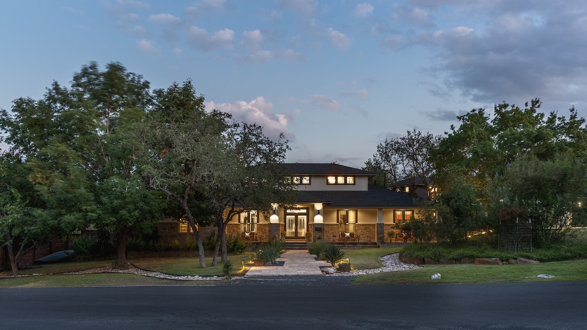 A contemporary retreat is visible from the street, showcasing a spacious front porch surrounded by landscaped greenery. The home features a combination of stone and modern siding, with multiple windows allowing natural light. Soft exterior lighting highlights the entry and garden areas as dusk approaches.