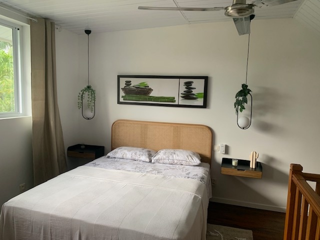 A cozy bedroom is presented with a queen-size bed featuring light bedding and a minimalist headboard. Wall-mounted shelves hold decorative items. Soft curtains frame a window, allowing natural light to illuminate the space. A ceiling fan offers airflow, enhancing comfort.