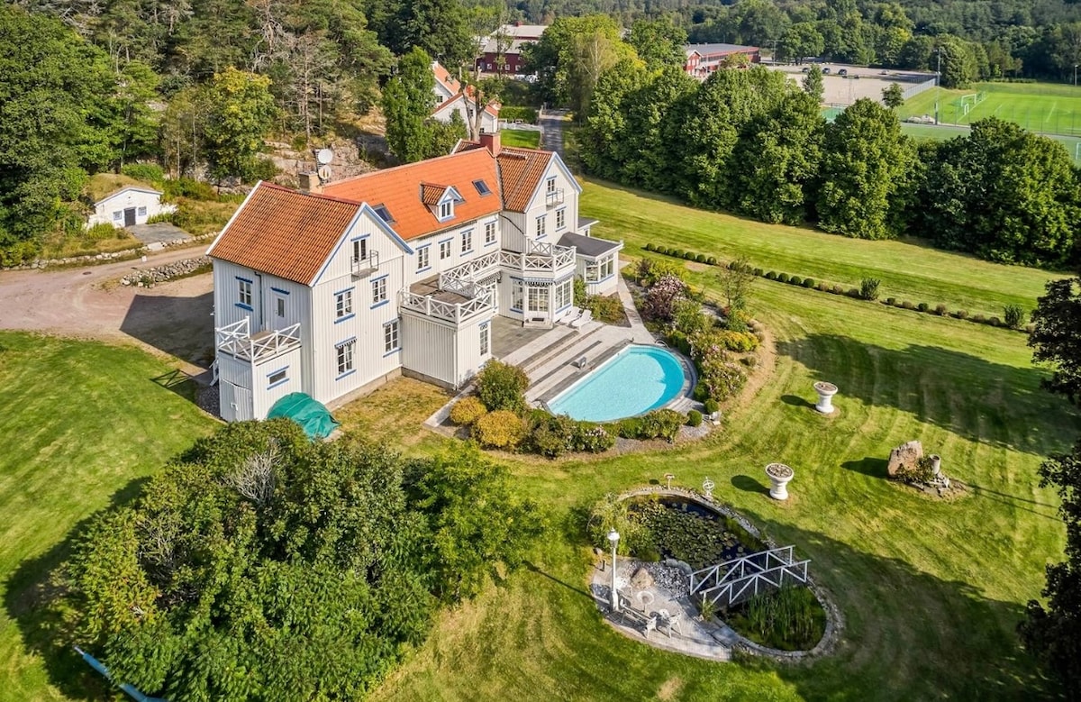 The property is set on lush green grounds, featuring a landscaped garden and a swimming pool. The facade of the mansion showcases multiple balconies and a charming roof, surrounded by peaceful nature. Nearby open areas are visible, contributing to the serene environment.