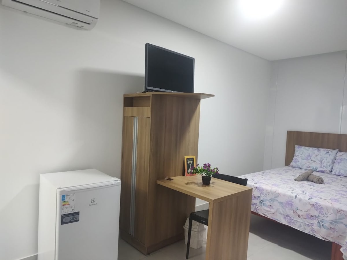 An organized room is featured with a spacious bed adorned in floral bedding. A small dining table with a chair is positioned beside a wooden cabinet, which holds a television. A compact refrigerator stands nearby, completing the functional living space.