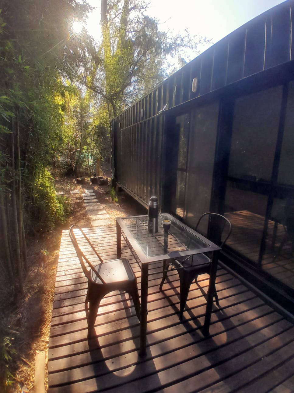 A wooden deck is seen outside a container structure, featuring a glass-top table with two metal chairs. Sunlight filters through surrounding trees, creating a natural ambiance in the tranquil setting.