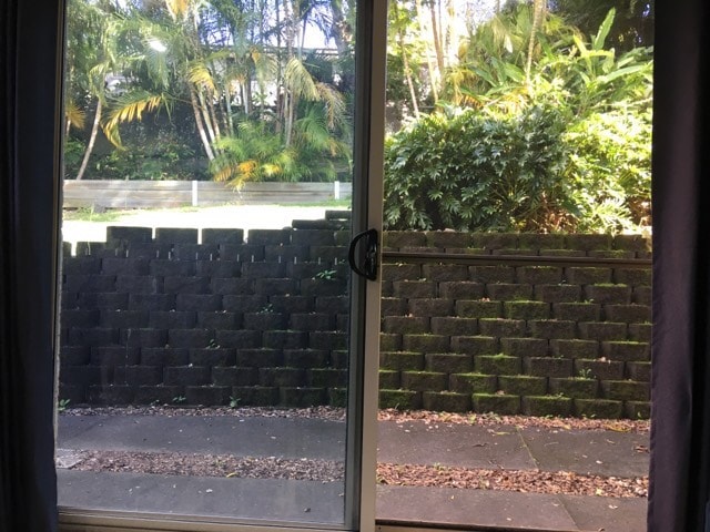 A sliding glass door reveals a view of a lush garden area, featuring tropical plants and a stone wall. The ground shows scattered leaves, and a walkway leads through the greenery, promoting a sense of tranquility and privacy.