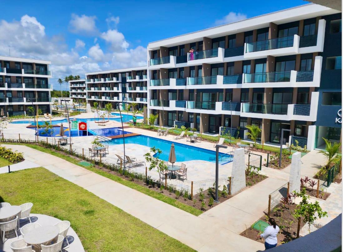An aerial view showcases multiple swimming pools surrounded by well-maintained gardens. Balconies of contemporary apartment buildings overlook the area, allowing for a peaceful retreat. Tables and chairs are arranged on a grassy area, providing space for relaxation and socializing.