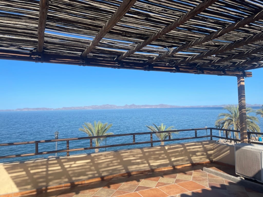 A shaded terrace overlooking the ocean features a wooden lattice roof. The panoramic view captures the calm blue waters and distant mountains. Palm trees line the edge of the deck, creating a serene outdoor setting.