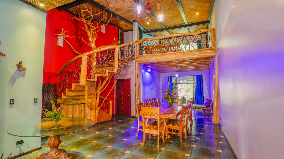 A spacious dining area features a large wooden table surrounded by matching chairs. A unique wooden staircase with a tree design leads to an upper level. Bright colors and artistic lighting enhance the modern design, while large windows allow natural light to fill the space.