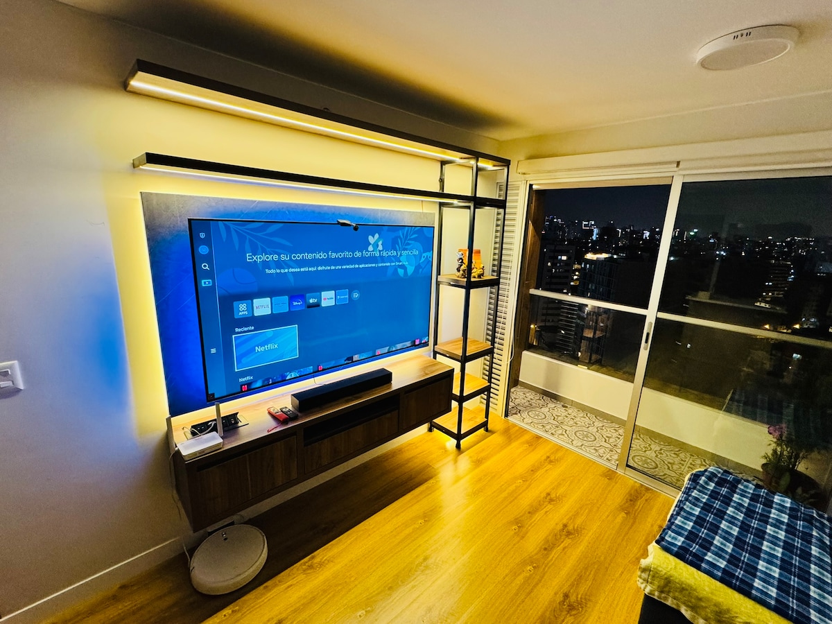 A modern living area is illuminated by soft lighting, featuring a large flat-screen TV mounted on the wall above a wooden media console. A sleek bookshelf stands beside the TV, and floor-to-ceiling windows provide views of the cityscape at night.