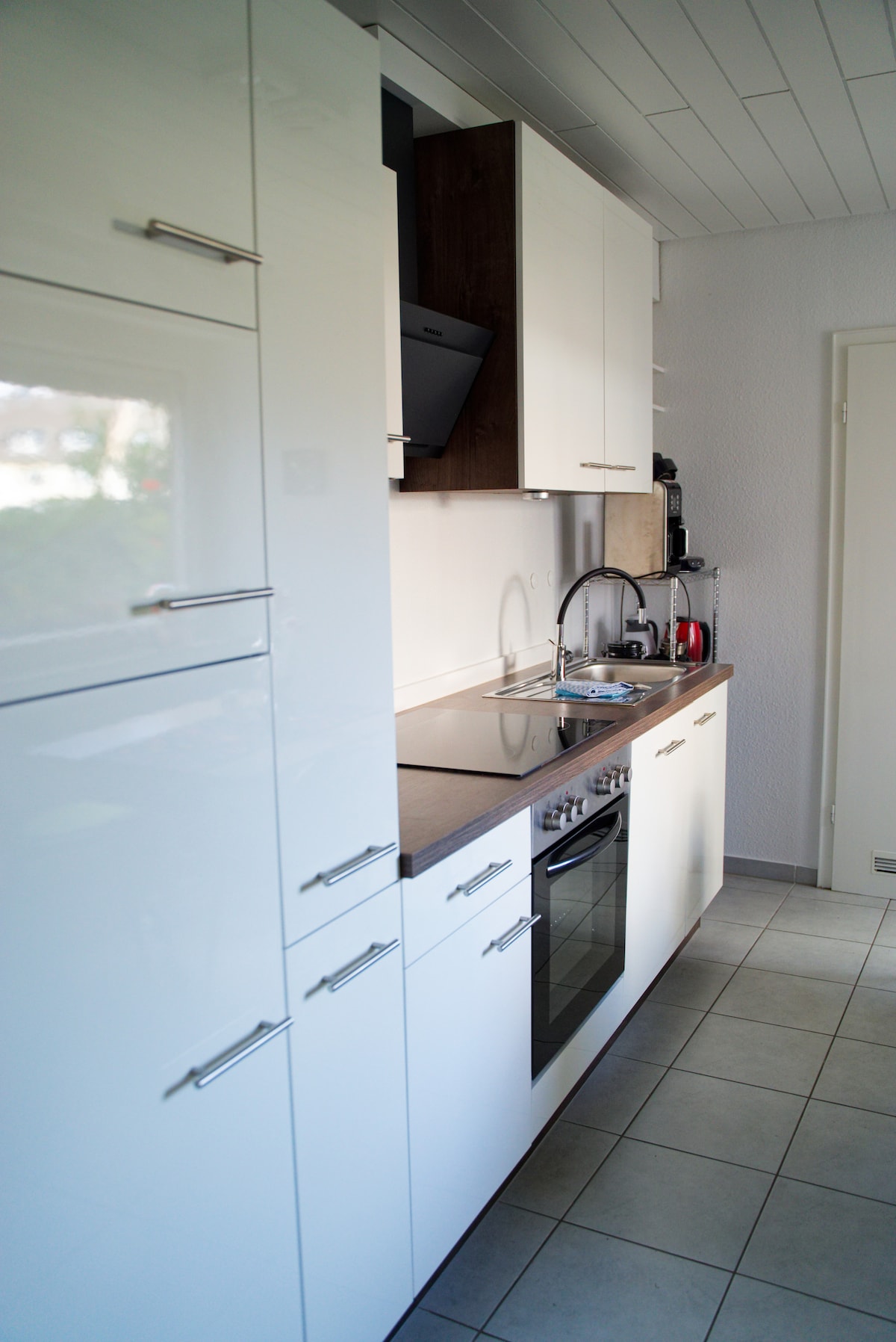 A modern kitchen features sleek white cabinetry paired with dark wood accents. Essential appliances, including an oven and a stovetop, are integrated seamlessly. Counter space is utilized efficiently, with a sink and coffee maker positioned for convenience. Light tiles provide a bright and clean environment.