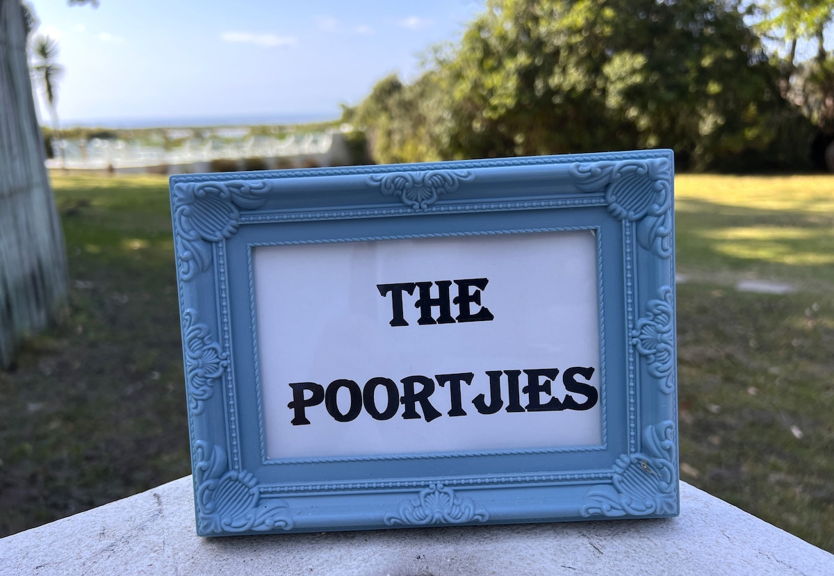 A decorative sign reading 'THE POORTJIES' is displayed in a light blue frame, set against a backdrop of greenery and a serene landscape. The horizon features glimpses of the lagoon and ocean, creating a calm atmosphere.