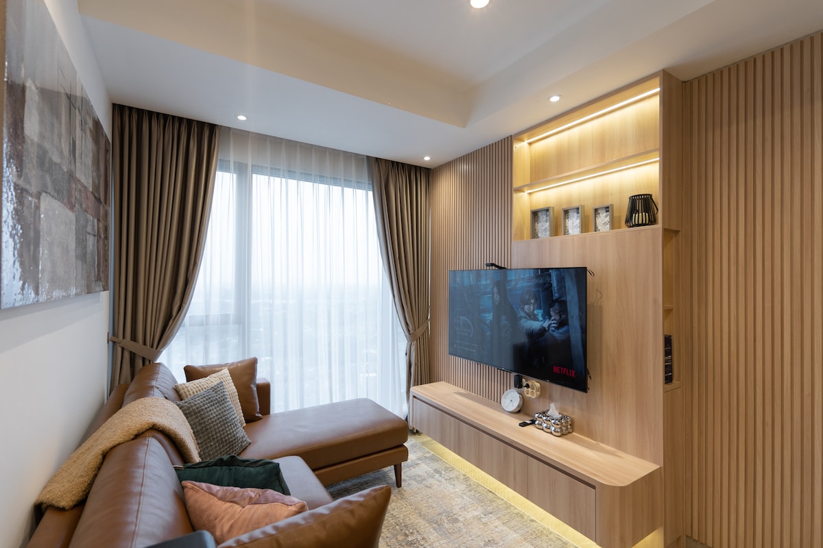 The living room showcases a cozy L-shaped sofa adorned with pillows and a blanket. A 55-inch Smart TV is mounted on a wooden entertainment unit, which is complemented by soft lighting. Floor-to-ceiling curtains frame a large window, adding warmth to the space.